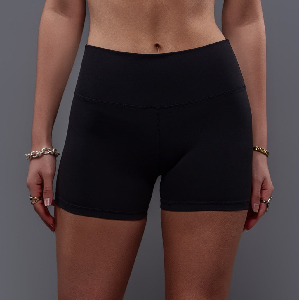 Someone wears Allure Black Shorts, made from stretchy black fabric with a flattering rise that highlights their midsection and upper thighs. Their ring- and bracelet-adorned hands rest at their sides against a plain gray background.