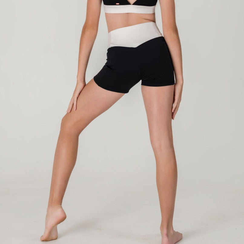 A person wears Chic Shorts - Charcoal- FINAL SALE and a black and white sports bra, standing barefoot with one leg bent and the other straight against a plain light background.