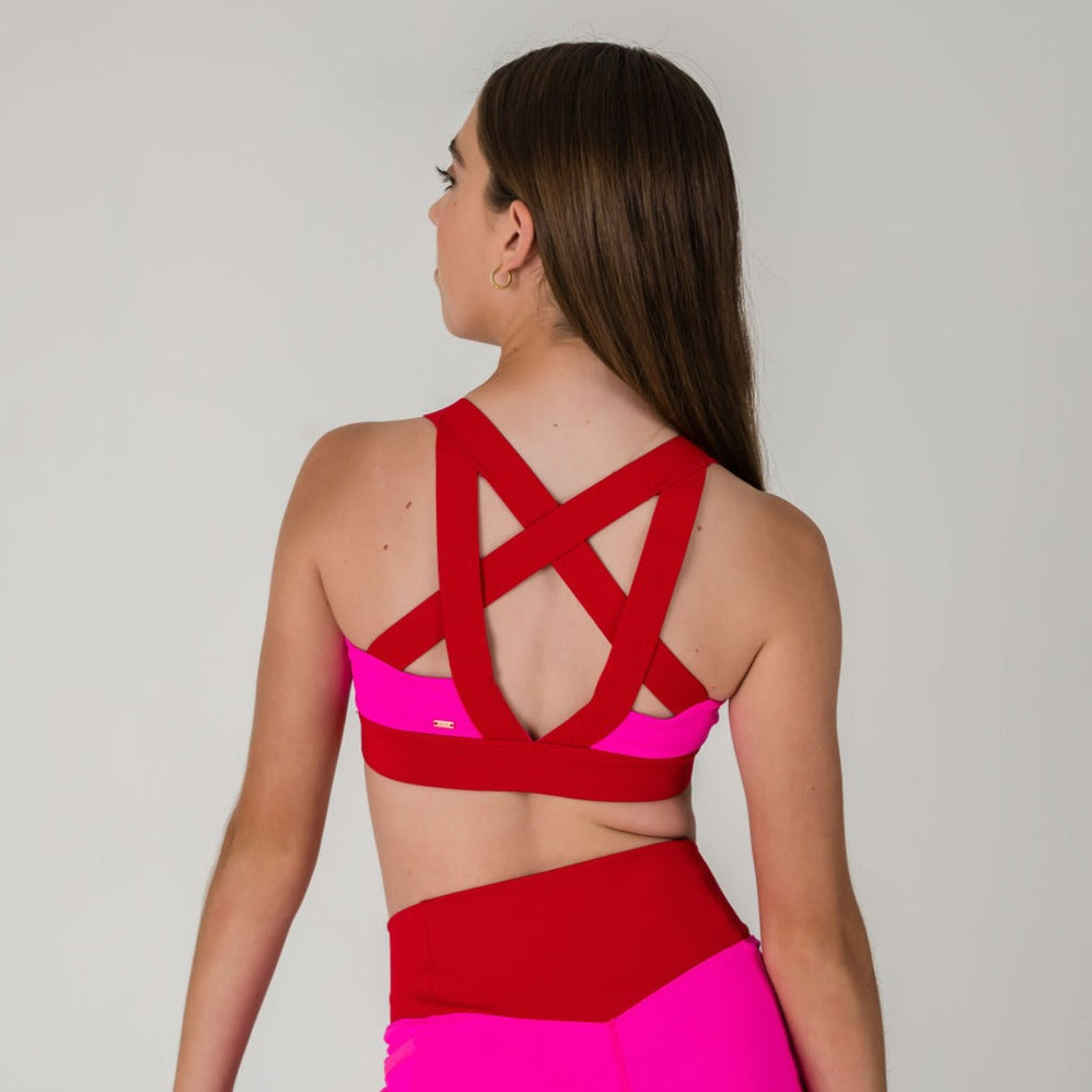 A woman with long brown hair is seen from the back, wearing the Chic Top in punch pink with crisscross straps and matching high-waisted leggings, creating a chic athleisure look against a plain light background.