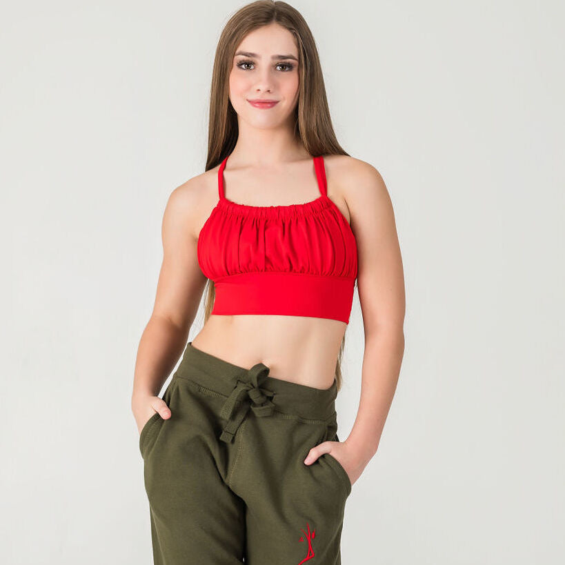 A young woman with long brown hair stands smiling against a plain light background, wearing the cherry red XOXO Top - FINAL SALE and high-waisted olive green pants, her hands in her pockets.