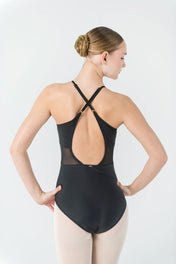A ballerina stands with her back to the camera, hands on hips, showing off the Ava Leotard's crisscross straps and open back. She pairs it with light-colored tights and wears her hair neatly in a bun.