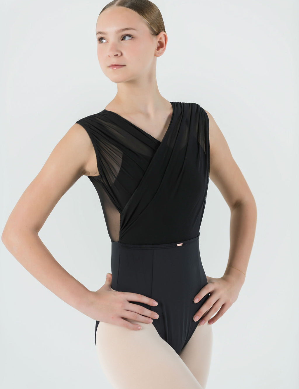 A young ballet dancer wears the Opera Leotard with crisscross mesh and light tights, standing hands on hips against a plain light background—a stylish choice for any dance wardrobe.