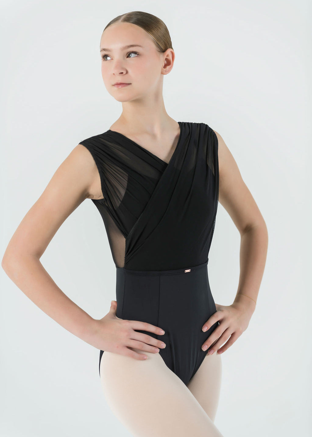 A young ballet dancer wears the Opera Leotard with crisscross mesh and light tights, standing hands on hips against a plain light background—a stylish choice for any dance wardrobe.