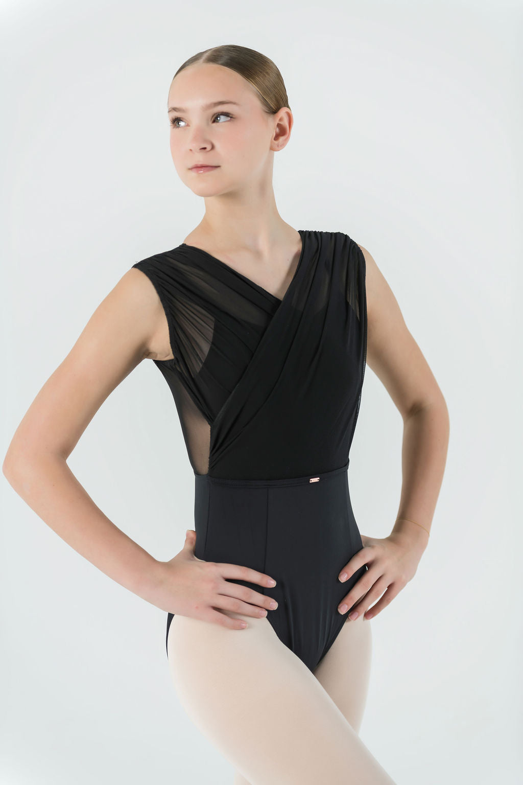 A young ballet dancer wears the Opera Leotard with crisscross mesh and light tights, standing hands on hips against a plain light background—a stylish choice for any dance wardrobe.