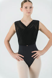 A young ballet dancer poses with hands on hips, wearing the Opera Leotard—featuring black crisscross mesh and sheer detailing—paired with pale tights against a light background. A chic addition to any dance wardrobe.