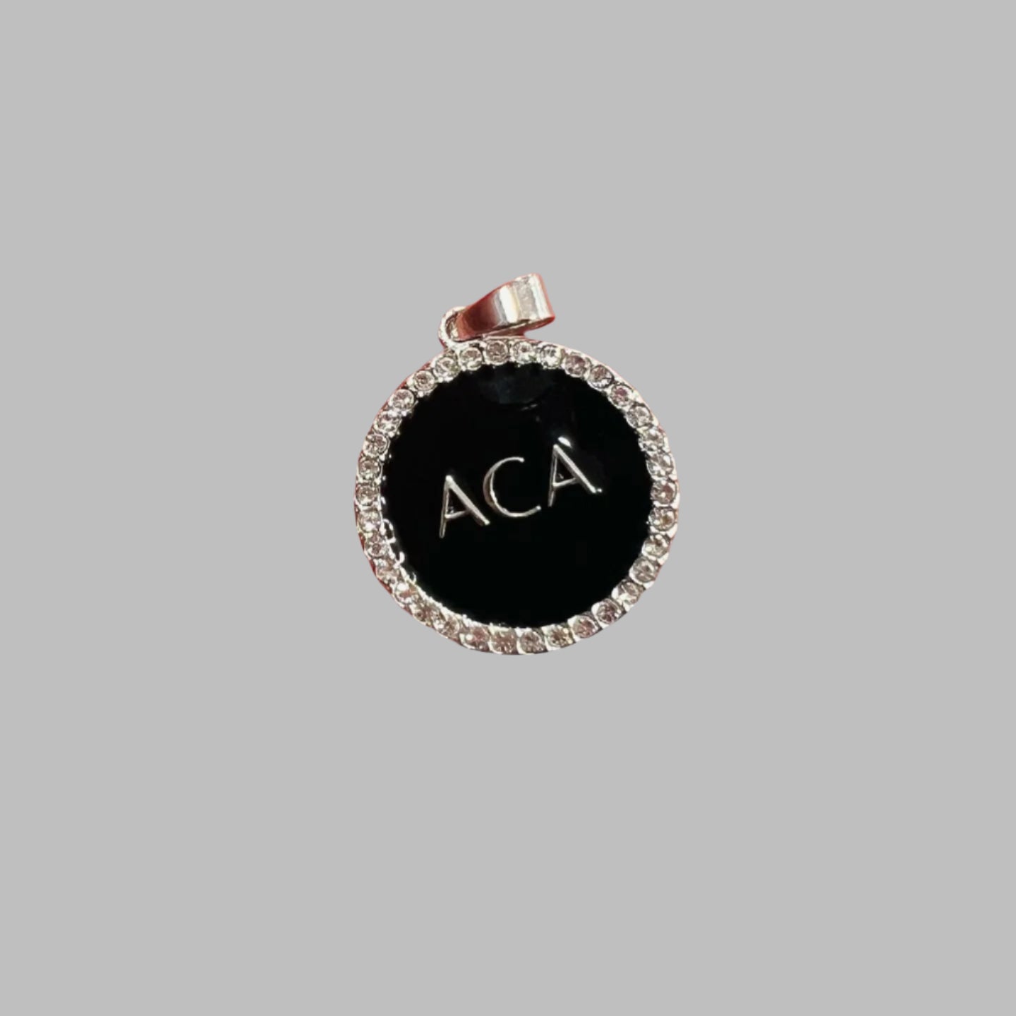 The Pendant Necklace - ACA features a round pendant with a black center, crystal charm border, and engraved ACA letters at the center, displayed on a plain gray background.