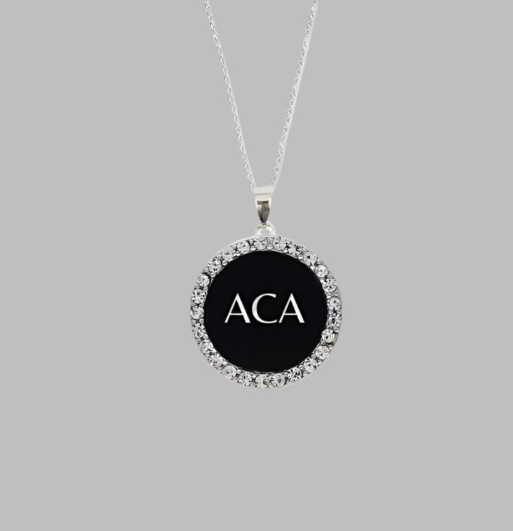 The Pendant Necklace - ACA is crafted in sterling silver with a round black center, clear crystal charm accents, and white ACA initials. It’s shown on a plain gray background for a timeless appeal.