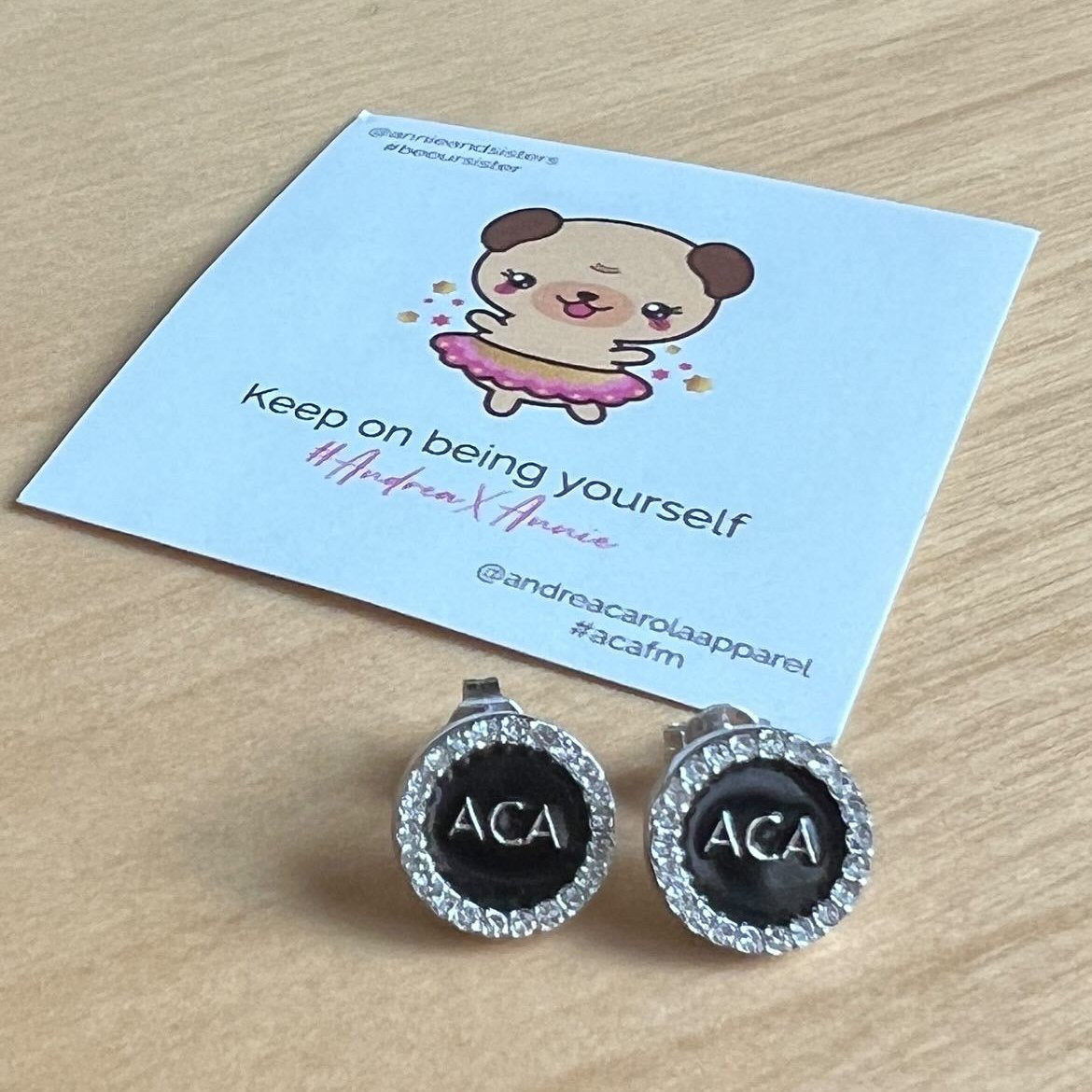 A pair of Stud Earrings - ACA, round with rhinestones and ACA in the center, rests on wood—evoking elegant sterling silver pave crystal style. Behind them is a card with a cute cartoon dog in a pink tutu and inspiring text.