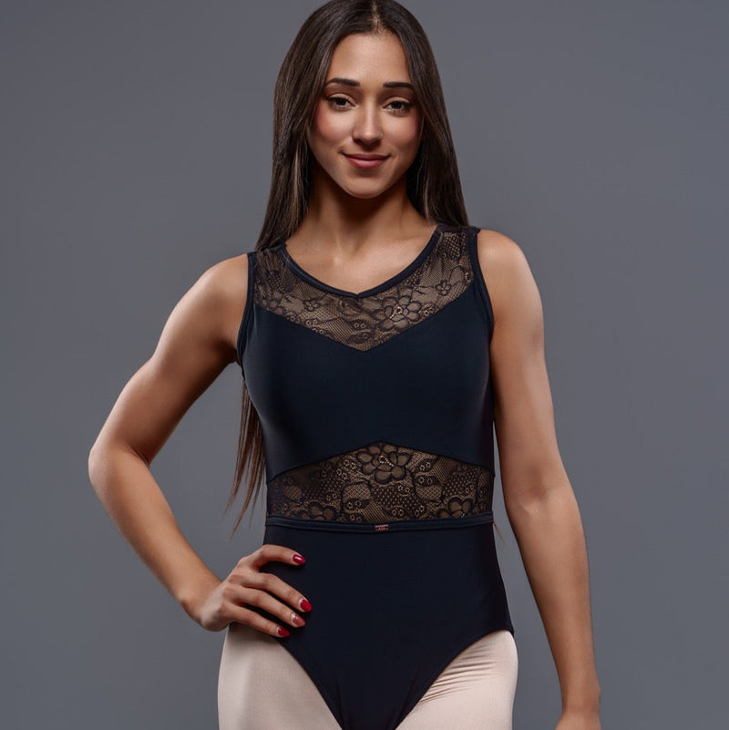 A woman with long brown hair stands confidently with one hand on her hip against a plain gray background, wearing the Lily Leotard—a black ballet piece with lace details—paired with beige tights.