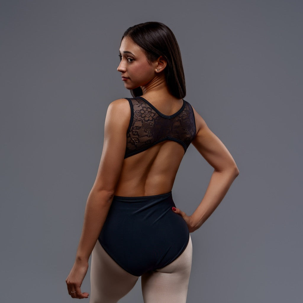 A woman models the Lily Leotard, a black ballet piece with floral lace details, paired with light tights as she stands facing away and looks over her shoulder against a plain gray background.