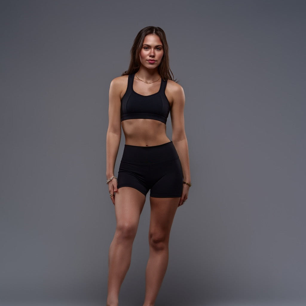 A woman modeling Allure Shorts - Black stands confidently against a plain gray background, one hand on her thigh and a relaxed expression, highlighting the flattering fit and wide waistband of these black athletic shorts.