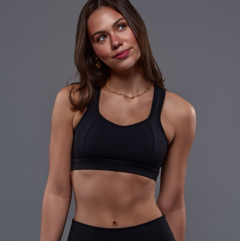 A young woman with long brown hair wears the Allure Top - Black with contrast stitching and black leggings, standing against a gray background and looking slightly to the side with a gentle smile.