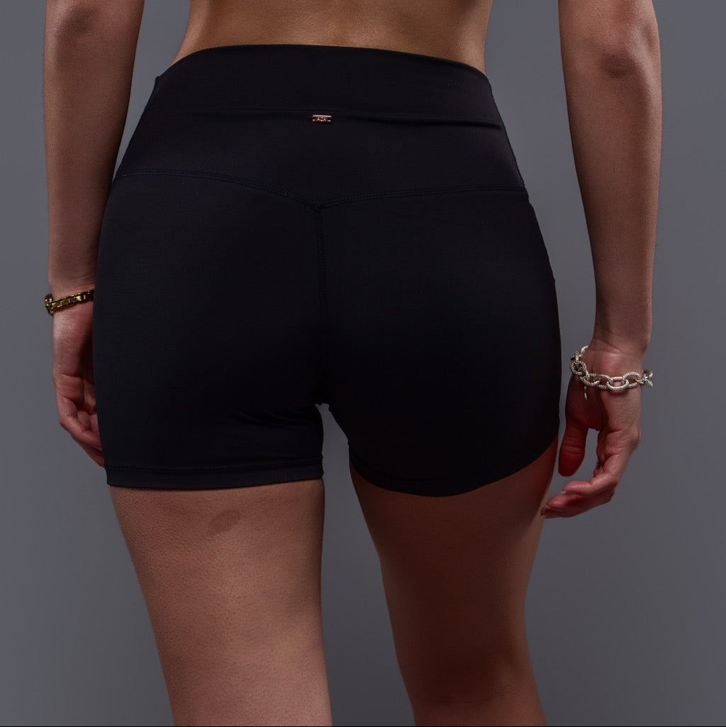 A person wearing Allure Shorts -Black with a wide waistband stands against a plain gray background, shown from the back. Bracelets on both wrists enhance the flattering fit.