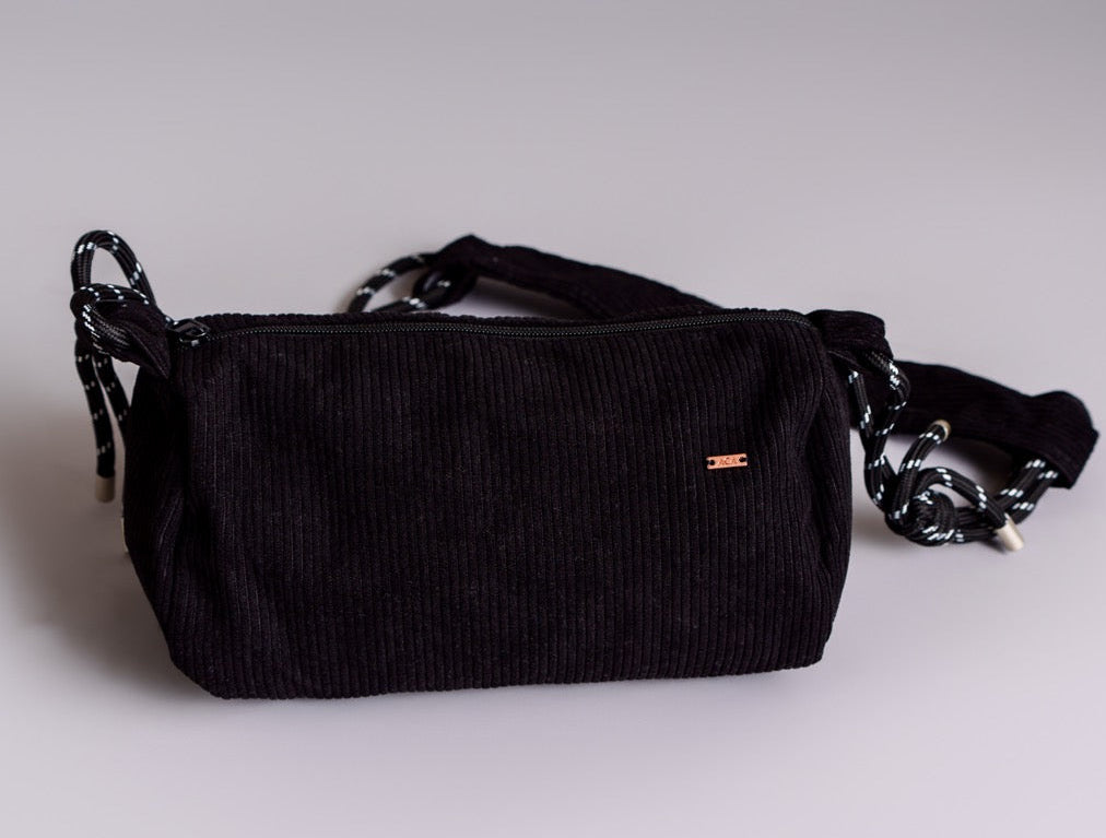 The Crossbody bag is a small black corduroy crossbody with a zip closure and a black-and-white striped adjustable strap, shown on a light gray background.