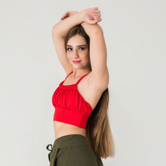 A young woman with long brown hair poses with her arms raised, wearing the XOXO Top - Cherry Red and olive green pants, standing against a white background and smiling gently at the camera.