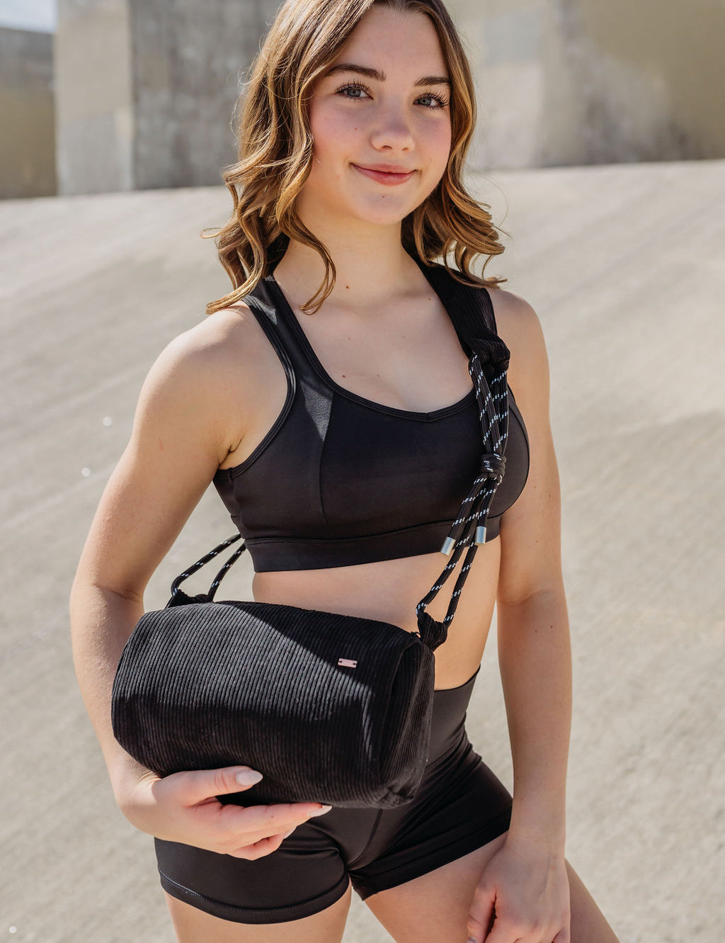 A young woman in a black sports bra and shorts stands outdoors, smiling at the camera while holding the Crossbody bag against a concrete background with sunlight casting shadows.