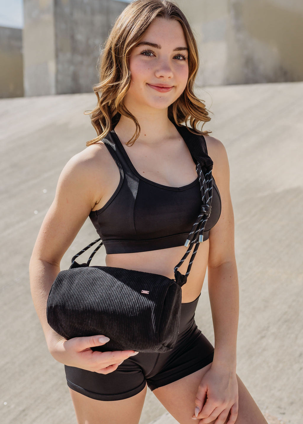 A young woman in a black sports bra and shorts stands outdoors, smiling at the camera while holding the Crossbody bag against a concrete background with sunlight casting shadows.