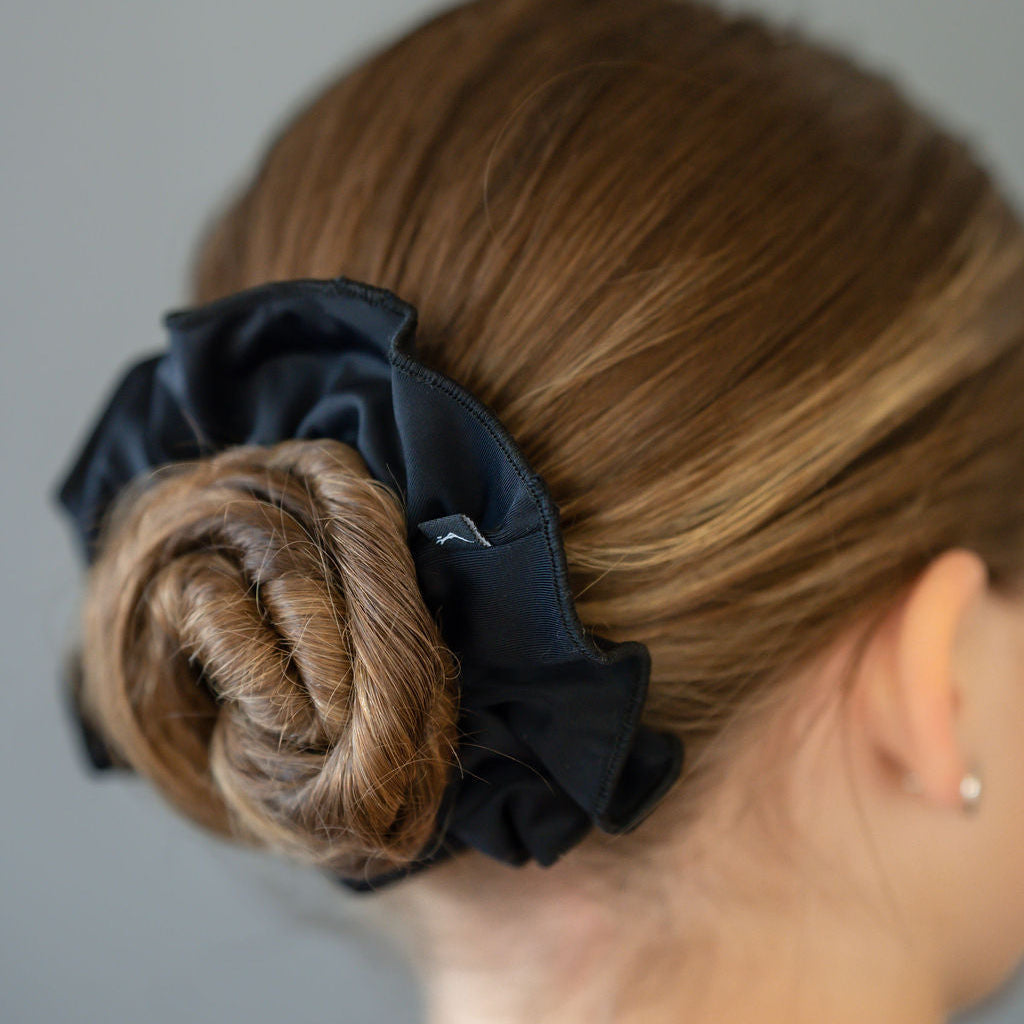 A woman with light skin wears her light brown hair in a low bun, secured by a large black scrunchie from the Scrunchies 2 Pack, demonstrating an elegant styling option.