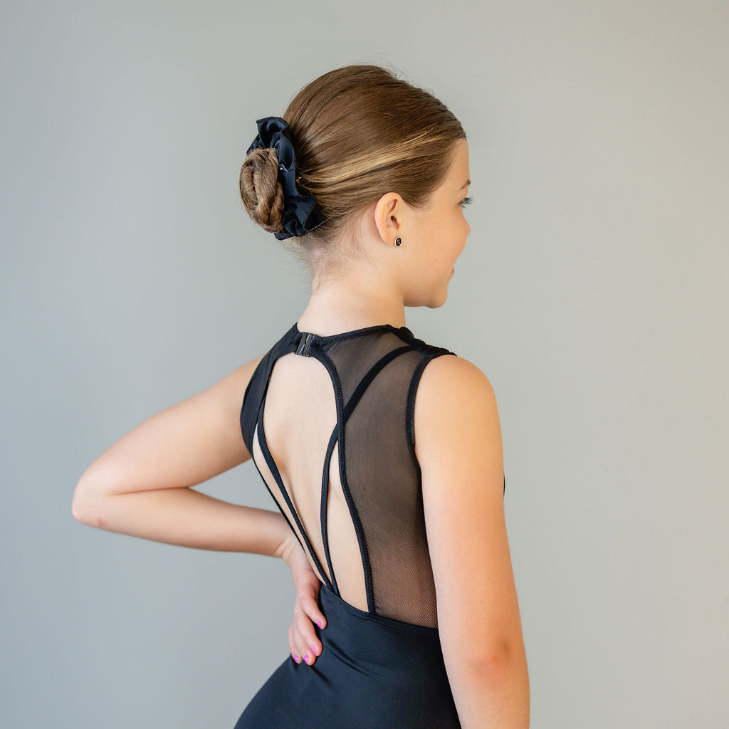 A young girl with light brown hair in a neat bun, secured with a Scrunchies 2 Pack, wears a sleeveless black leotard with sheer back and decorative straps. She stands sideways, hand on hip, against a plain background.