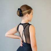 A young girl with light brown hair in a neat bun, secured with a Scrunchies 2 Pack, wears a sleeveless black leotard with sheer back and decorative straps. She stands sideways, hand on hip, against a plain background.