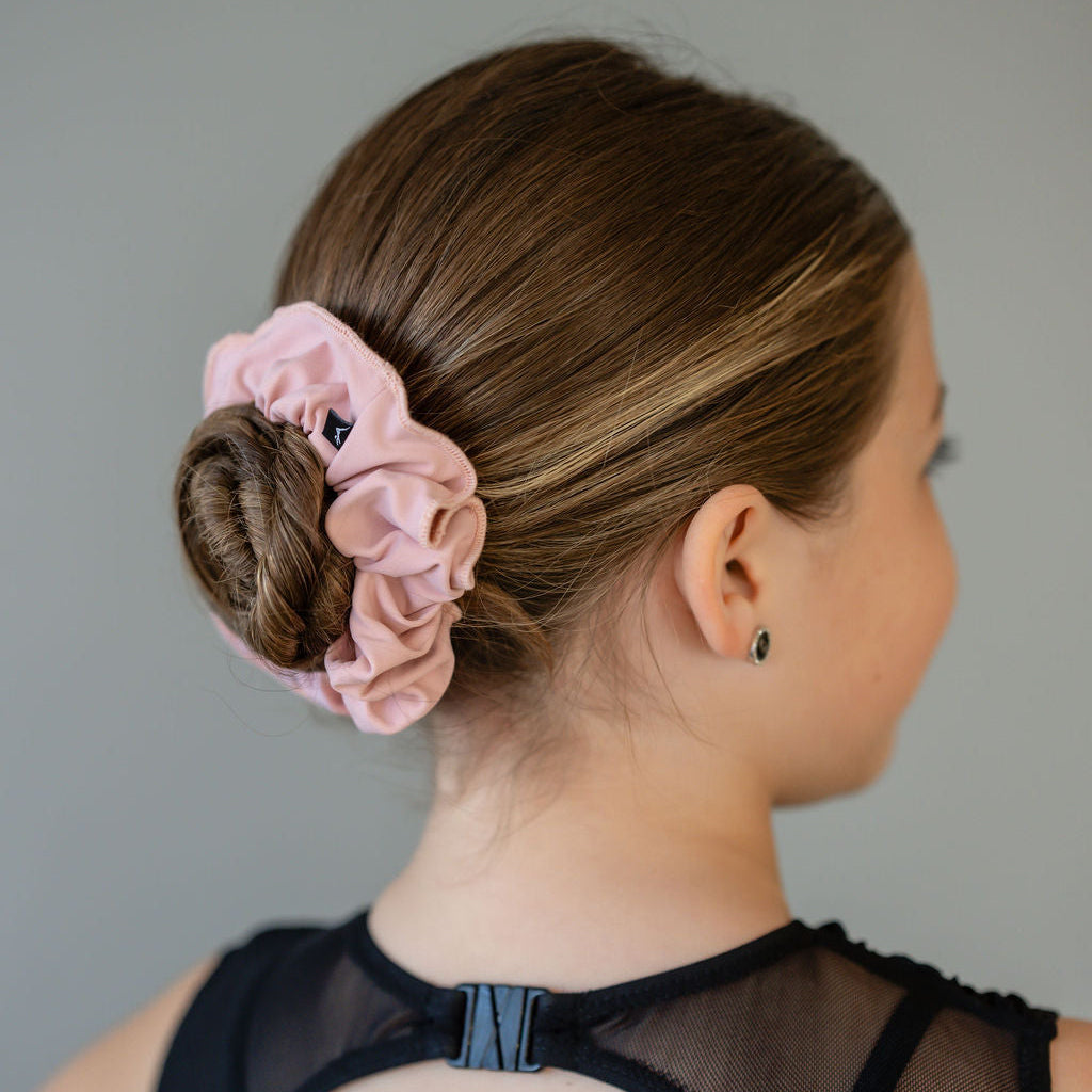 A young girl with brown hair in a bun, styled with a light pink Scrunchies 2 Pack hair tie. She wears a black top with a sheer mesh back and black stud earrings. The pink scrunchie adds softness to the neutral background.