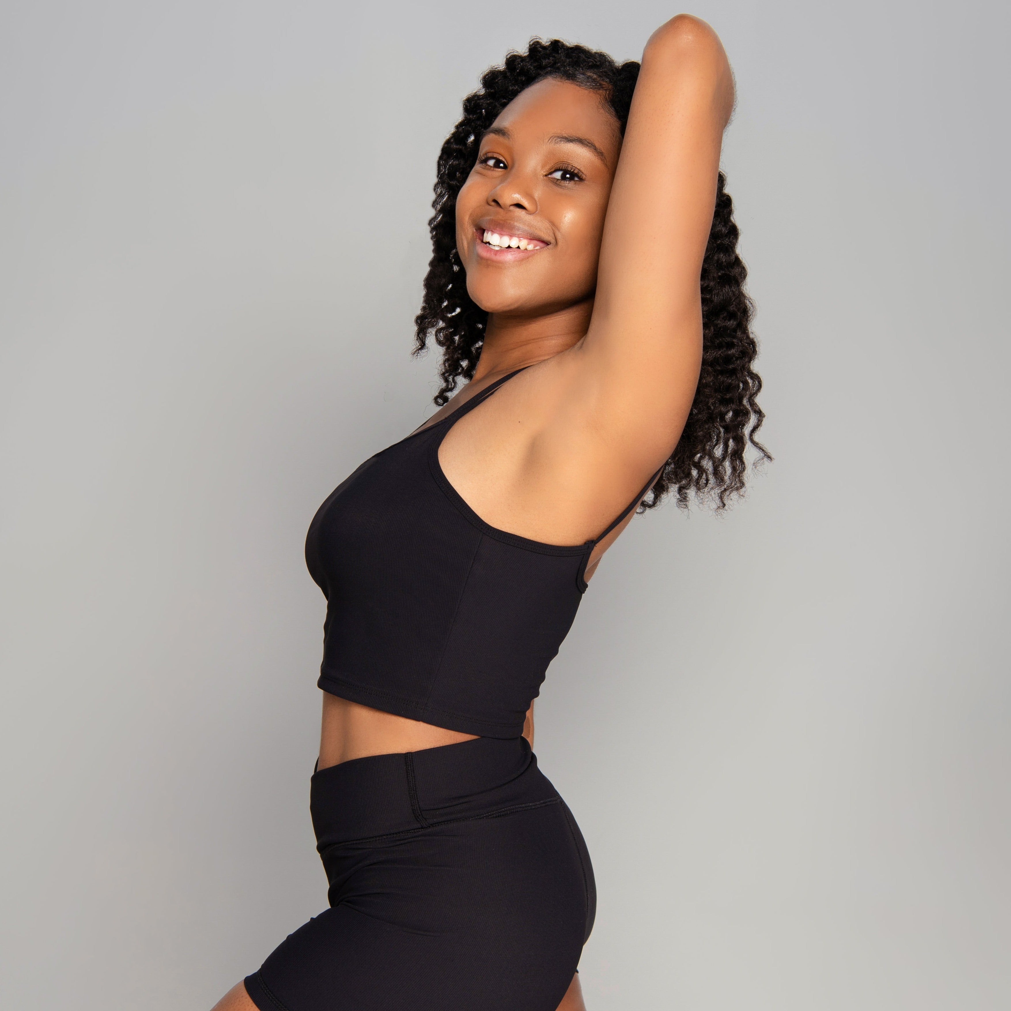 A woman with curly hair smiles confidently, wearing an ICONIC2 Tank with removable pads and matching black shorts, posing with one arm raised behind her head against a plain gray background.