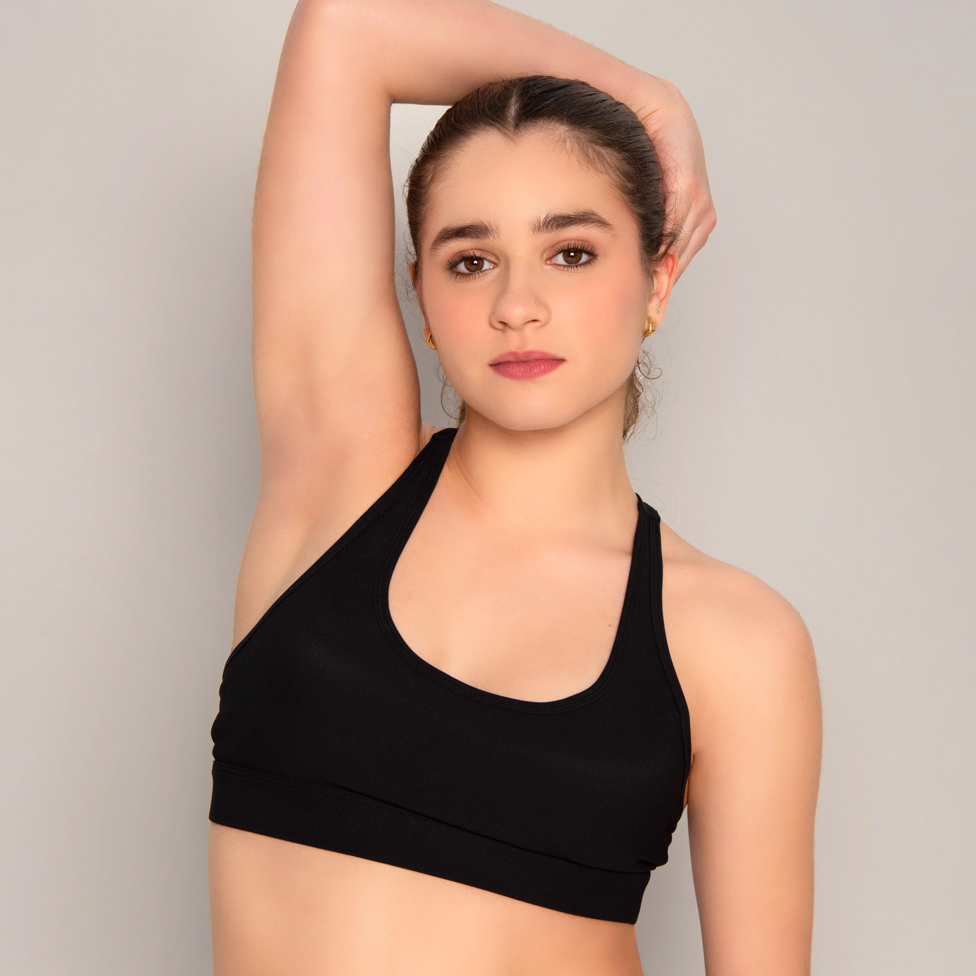 A young woman models the 2Iconic Sport Bra in black, standing against a neutral background with one arm raised, exuding confidence and showcasing this modern athletic essential.