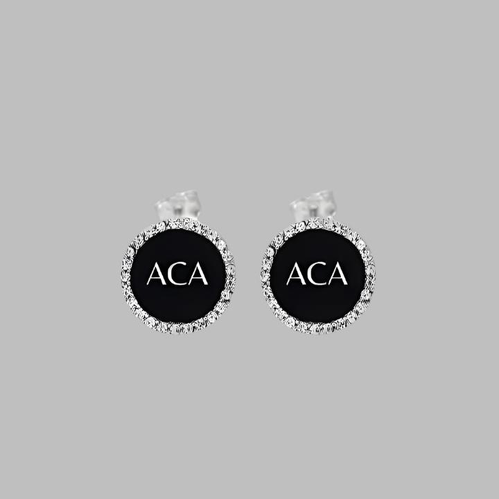 A pair of Stud Earrings - ACA, round and black with white letters at the center surrounded by small clear gemstones, are shown on a plain gray background.