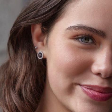 Close-up of a young woman with light skin and brown hair, revealing one green eye and part of a smile. She wears Stud Earrings - ACA—one features black enamel, the other is a delicate sterling silver pavé crystal stud above it.
