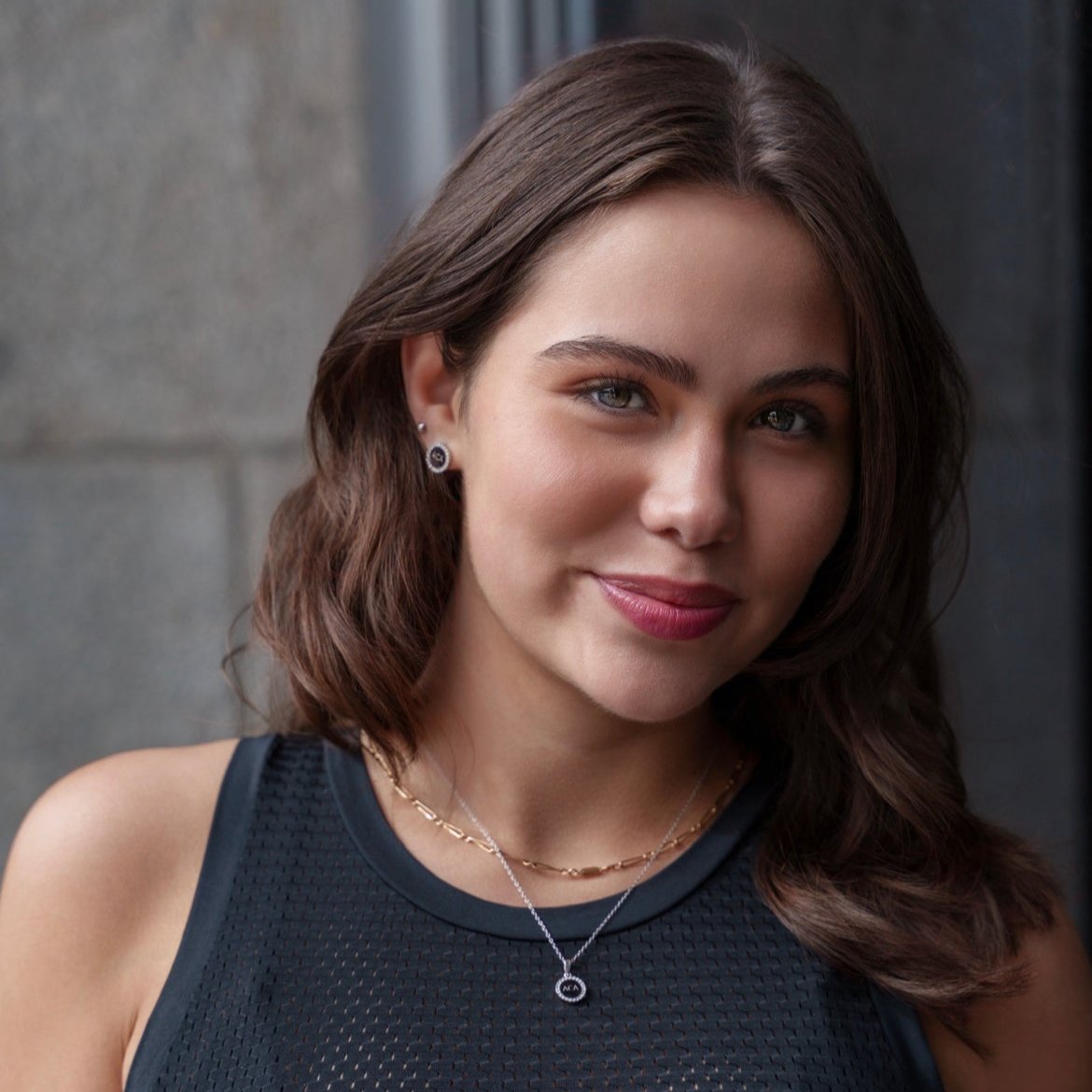 A young woman with wavy brown hair and blue-green eyes smiles softly, wearing a sleeveless black top, simple jewelry, and the Pendant Necklace - ACA as she stands before a gray stone wall.