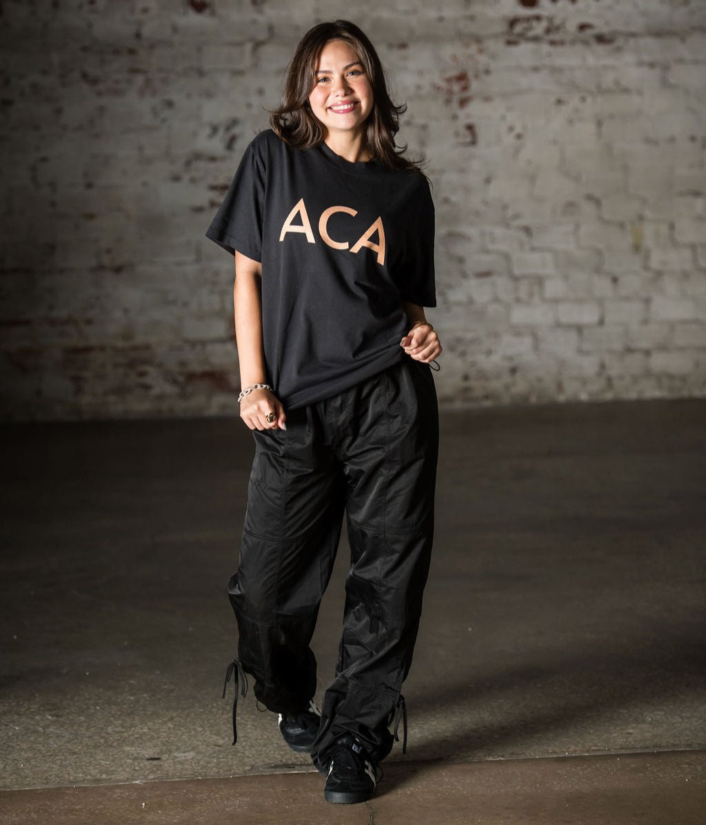 A smiling woman stands indoors before a white brick wall, wearing the Oversized T-shirt with a faded cotton feel and large tan ACA logo, paired with black pants and shoes. She poses, holding her shirt with one hand.