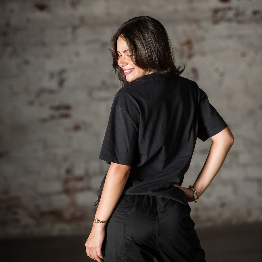 A woman with shoulder-length brown hair smiles over her shoulder while modeling the Oversized T-shirt and black pants, standing sideways in front of a textured, light-colored brick wall.
