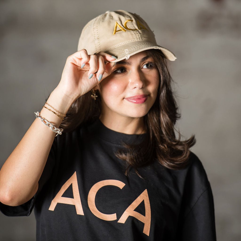 A woman with long brown hair, wearing a Lightly Distressed Cap with an embroidered ACA logo and a black t-shirt reading ACA in bold letters, smiles gently while holding the brim of her cap. The softly blurred background adds warmth to the scene.
