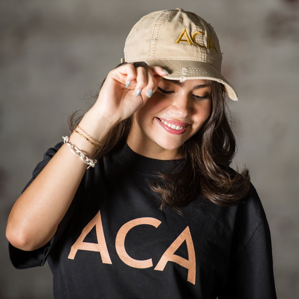 A smiling woman with long dark hair wears the Lightly Distressed Cap in beige and a black shirt, both displaying the bold embroidered ACA logo. She tilts her cap forward with one hand.
