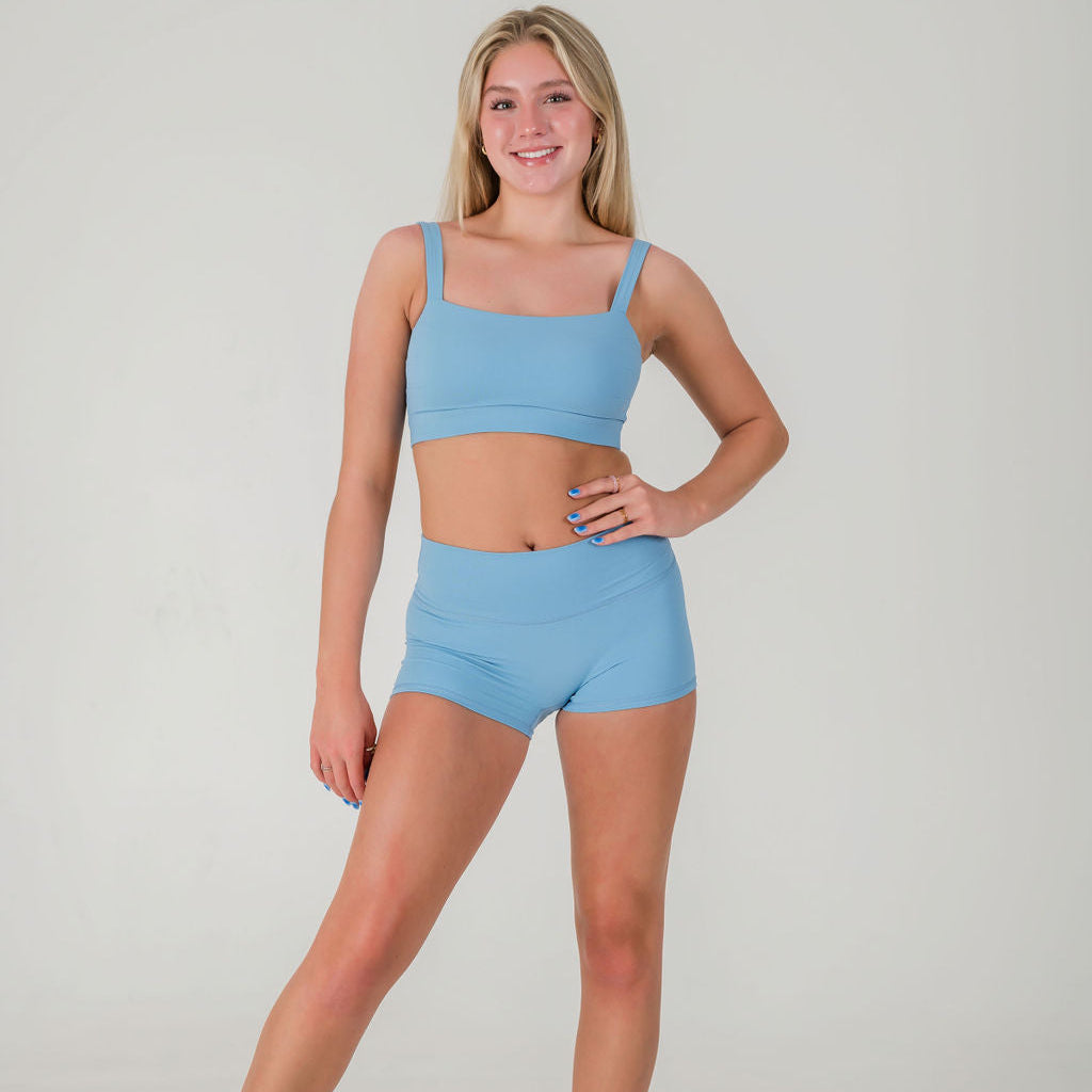 A young woman with long blonde hair stands smiling against a plain background, wearing FRESH Shorts in sky blue and a matching sports bra. One hand rests on her hip while the other hangs naturally by her side.