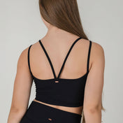 A woman with long brown hair, seen from the back, is wearing the ICONIC2 Tank in black ribbed fabric with double straps and matching black bottoms, set against a simple light background.