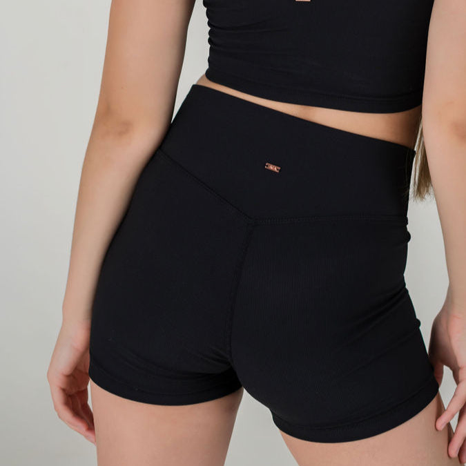 Model shown from the back wears 2Iconic Shorts in black with a V-front waist and ribbed texture, paired with a black crop top, left arm relaxed at their side against a plain light background for a flattering fit.