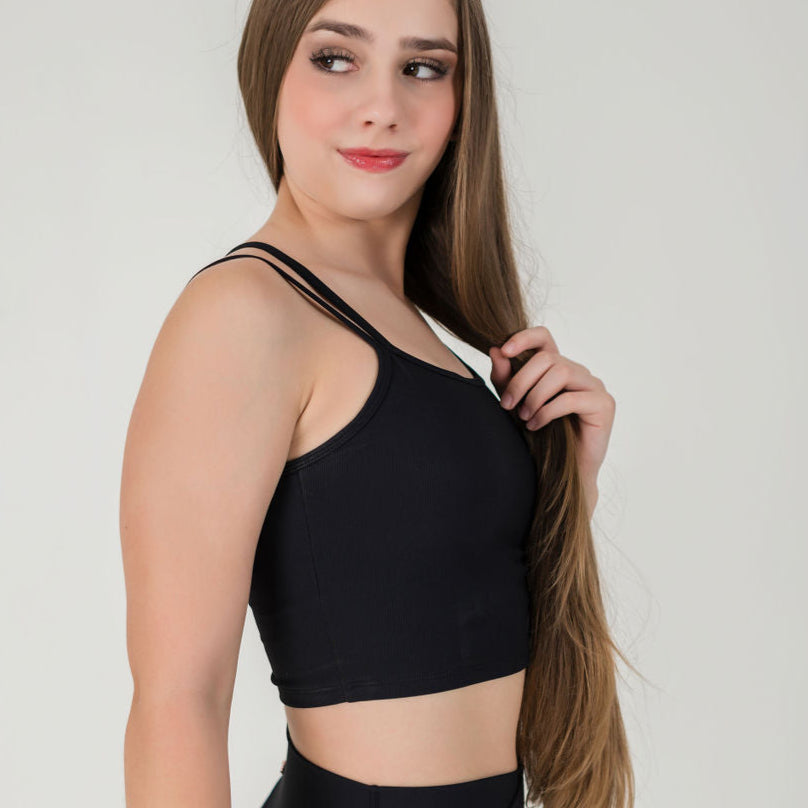 A woman with long brown hair wears the ICONIC2 Tank and black high-waisted bottoms, standing sideways as she gently holds her hair and smiles against a plain light background.