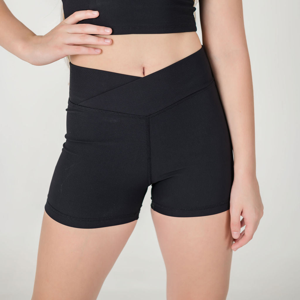 Wearing ICONIC2 Shorts and a black crop top, a person stands against a plain light background. The image shows the torso and upper legs, highlighting ICONIC2 Shorts as must-haves for versatile, active style.