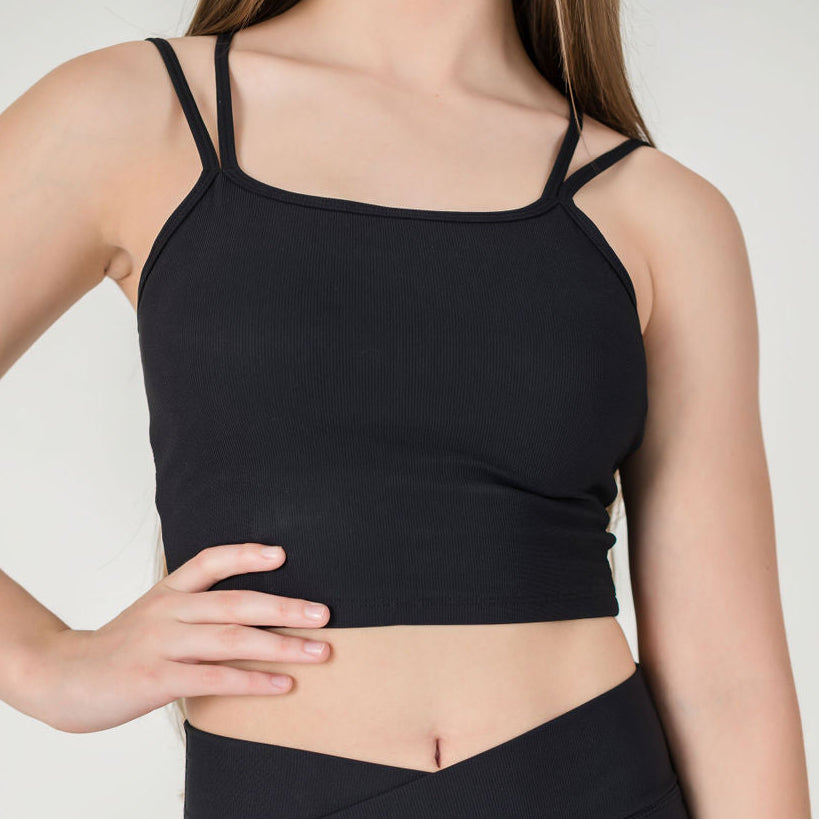 A woman with long brown hair wears the fitted black ICONIC2 Tank with double spaghetti straps and high-waisted black leggings, posing with one hand on her waist against a plain light background.