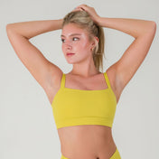 A woman wears the FRESH Top in lime yellow, standing against a plain background with arms raised and looking to the side. The photo highlights the square neck design and buttery soft fabric of this activewear top.