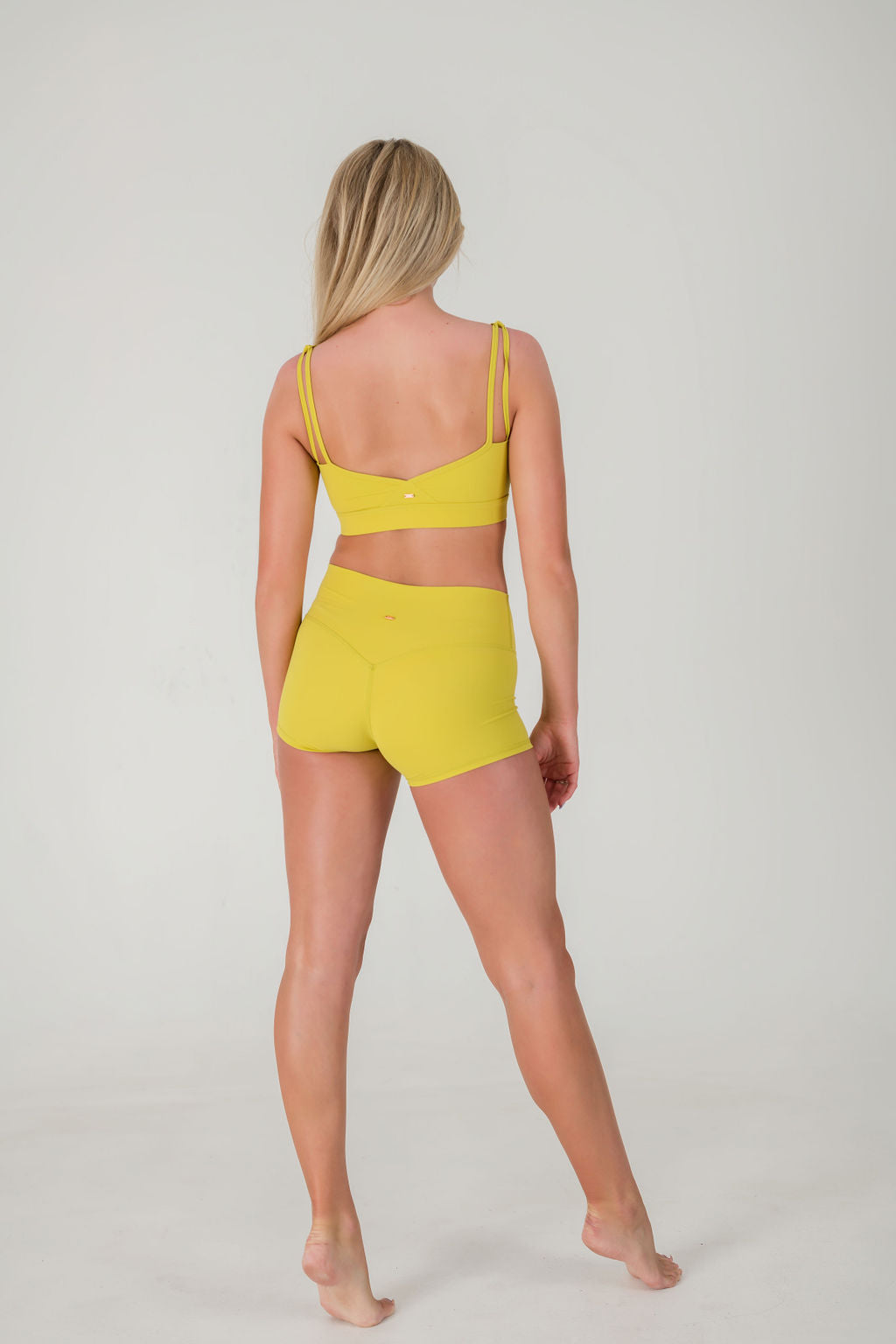 A woman with long blonde hair faces away, wearing the FRESH Top in lime yellow made from buttery soft fabric, paired with matching shorts against a plain white background.