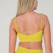 A woman with long blonde hair is seen from behind, wearing the FRESH Top in lime yellow, made from buttery soft fabric, against a plain light background.