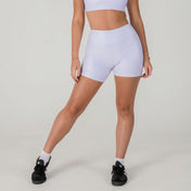 A woman stands with one leg forward against a plain light background, wearing Airsoft Shorts- Lavender Lux and a matching sports bra. She pairs the set with black sneakers and white socks; her head is not visible.