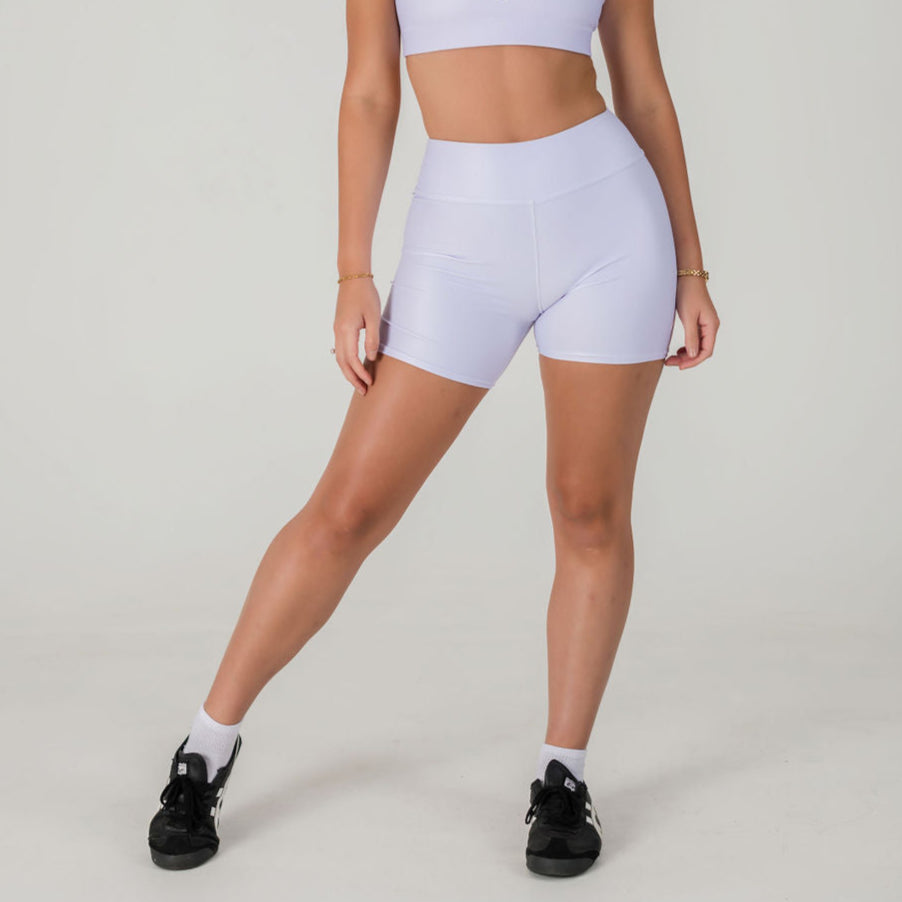 A woman stands with one leg forward against a plain light background, wearing Airsoft Shorts- Lavender Lux and a matching sports bra. She pairs the set with black sneakers and white socks; her head is not visible.