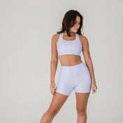 A woman stands against a plain light background wearing the Airsoft Sports Top - Lavender Lux with matching high-waisted shorts, offering maximum support. She looks to her left, one hand resting on her thigh.