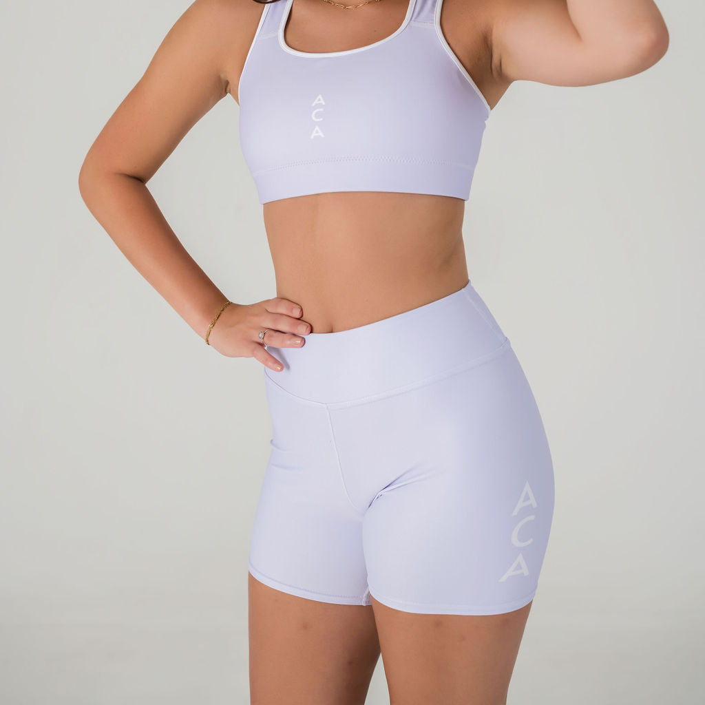 A person models the Airsoft Shorts–Lavender Lux, the ACA logo shows on the side, paired with a matching sports bra with ACA branding. Both pieces are shown against a plain light background; the persons face is not visible.