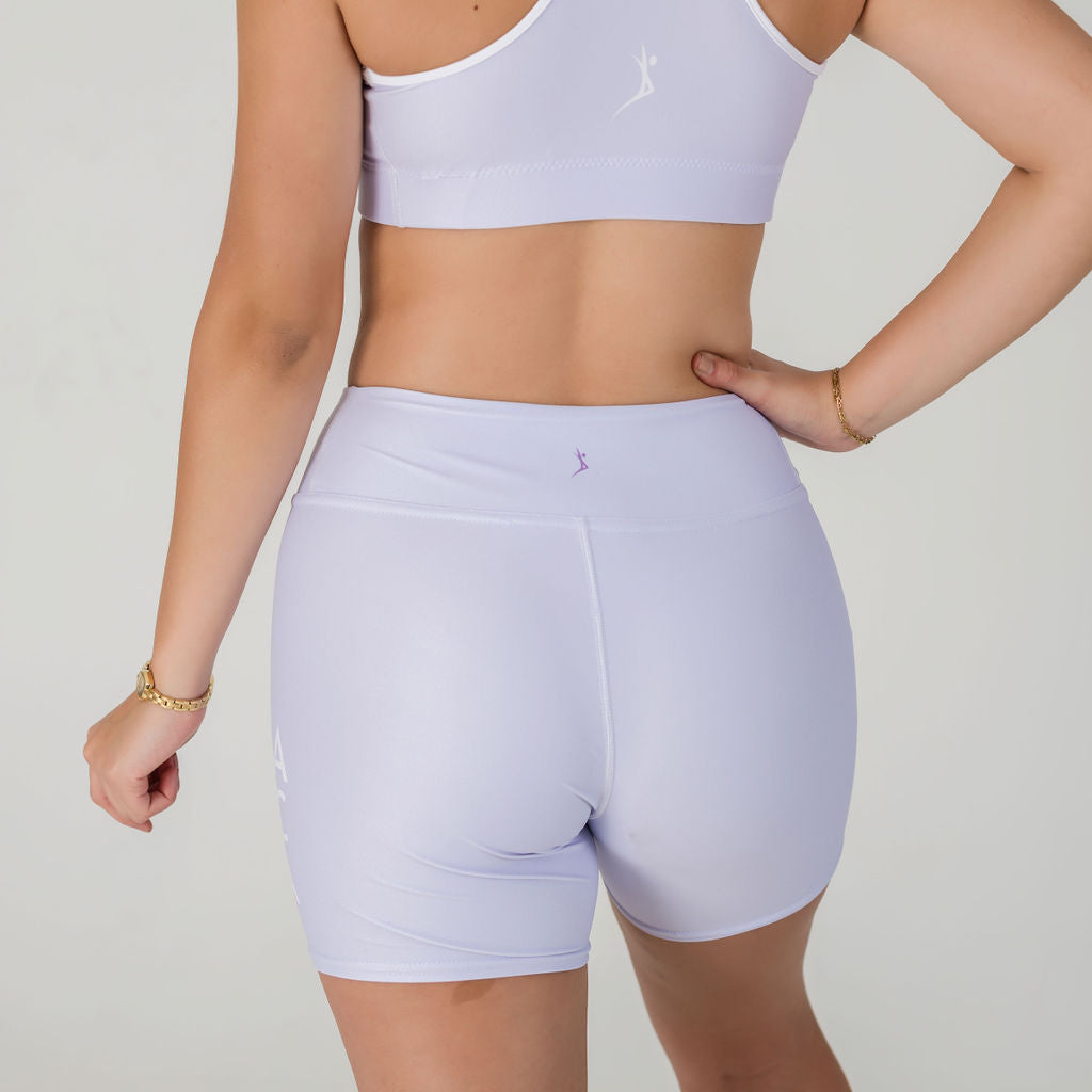 A woman stands with her back to the camera, wearing Airsoft Shorts- Lavender Lux and a matching sports bra, showing the outfit’s fit. Her left hand rests on her hip against a plain light background.