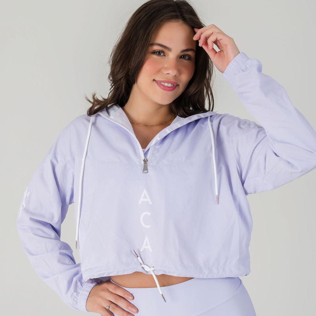 A woman with medium-length brown hair, smiling and touching her head, wears the Airsoft Anorak - Lavender Lux with breathable mesh lining and matching pants against a plain light background.