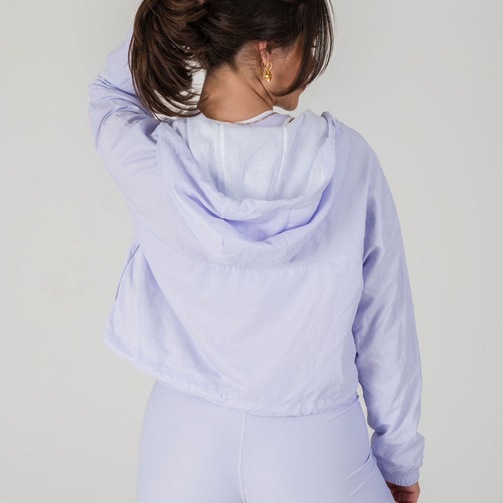 A woman with brown hair in a ponytail, wearing the Lavender Lux Airsoft Anorak and leggings, stands facing away from the camera against a plain light background.